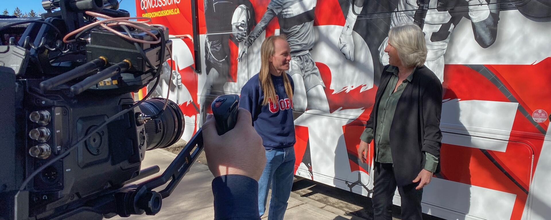 Man and a woman stand in front of a large bus, with a camera man in the foreground, filming them