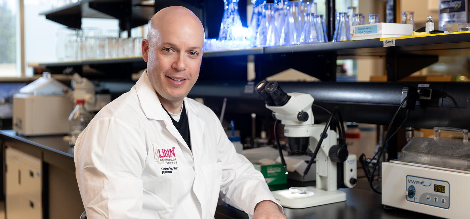 A man sits at a table with a microscope with a white lab coat on smiling at the camera