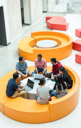 Students sit together in a circle with notebooks and laptops, deep in discussion.