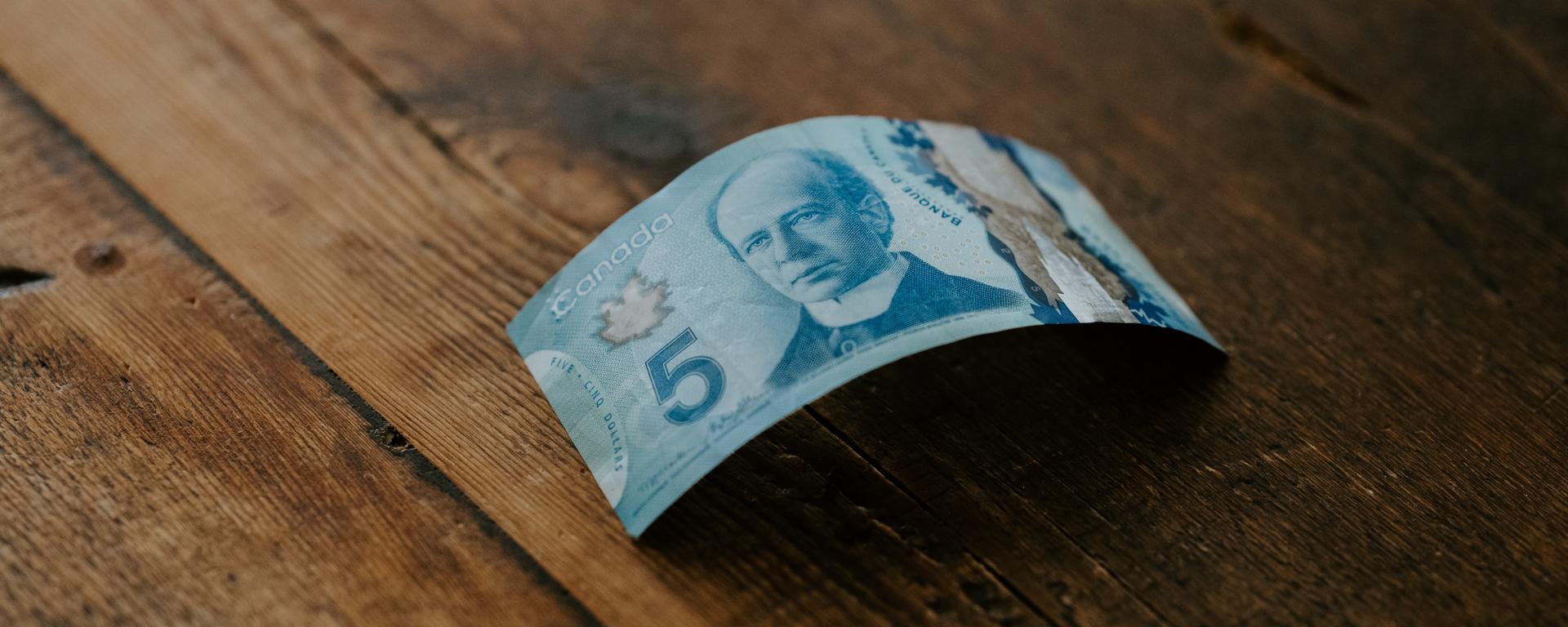 A Canadian five dollar bill sits on a wood table