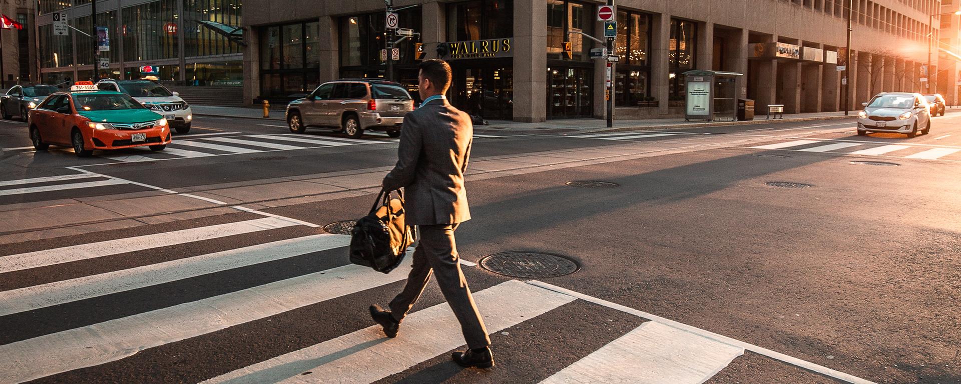 A person in a suit walks through a crosswalk on a sunny morning in a downtown city.