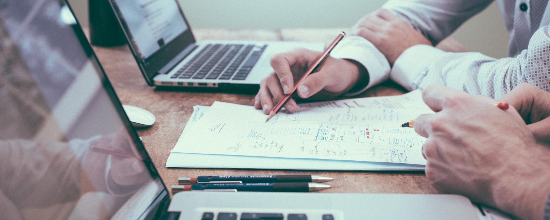 Two people sit at a wood desk. They each have a laptop in front of them. Between them is a stack of papers, and one person is point at the papers with a pencil in their hand.