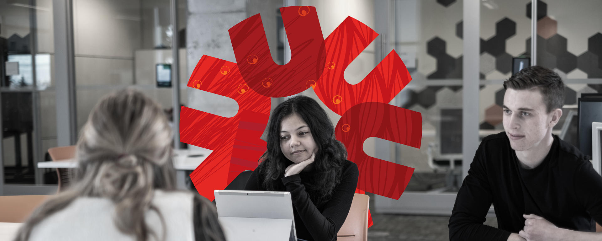 Three students sit at a table. One is looking at a tablet, one has a binder open in front of them, the third is facing the other two in conversation. 