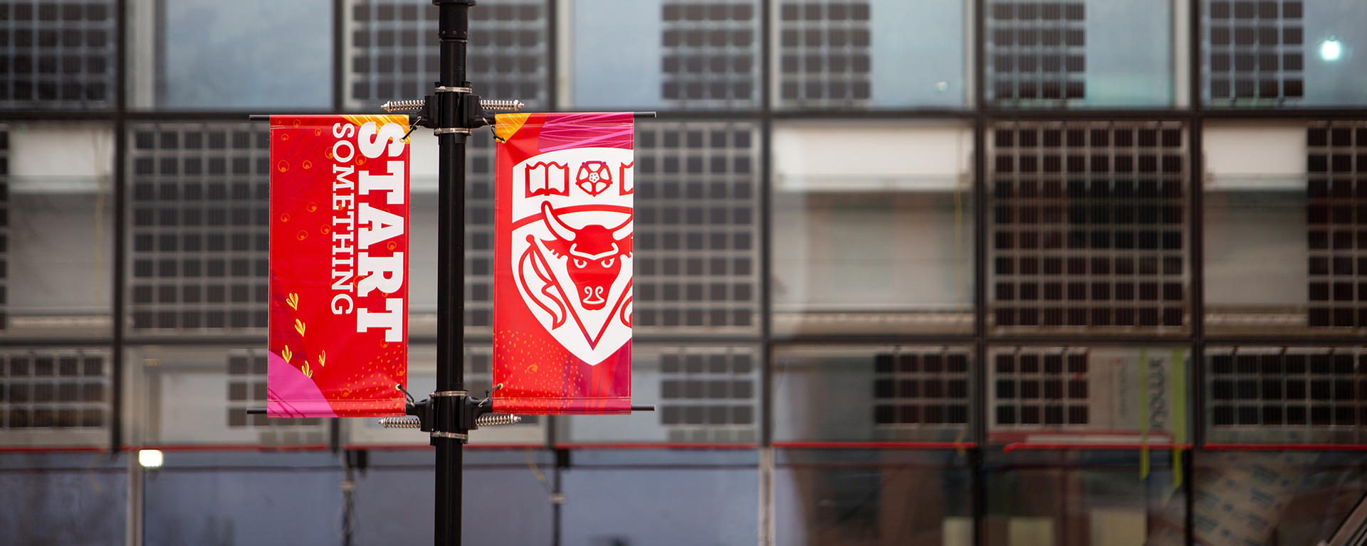 Two banners on a light standard in front of the building. One banner reads "Start Something" and the other has the UCalgary Coat of Arms.