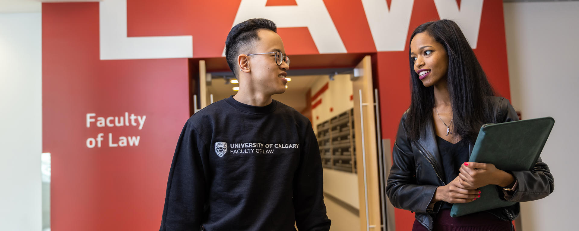 Two students walk through a hallway. They are carrying laptops and there is a red wall with "Faculty of Law" behind them.