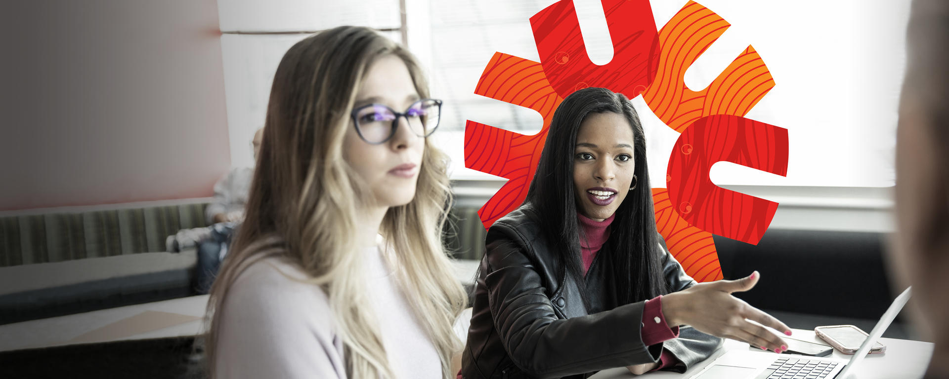 Two students sit at a desk talking to a third student