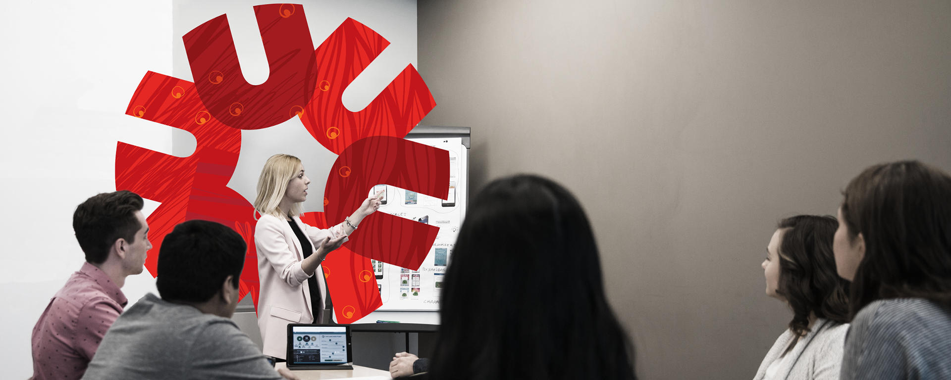 An instructor stands at the front of a room, gesturing to a large chart. 5 students sit in front of her facing her.