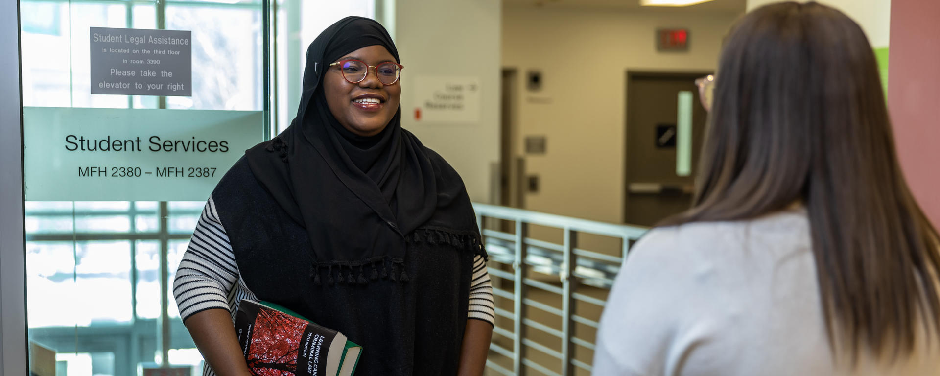 A student wearing a hijab stands in a hallway holding books and talking to another student. 