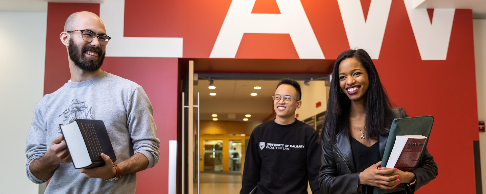 Three students walk through a doorway. The person on the left wears a grey sweatshirt and is carrying a textbook. The person in the middle is wearing a black sweatshirt with UCalgary Law on it. The person on the right has long dark hair and is carrying a laptop and a textbook. Behind them, the word "Law" is painted in large white letters over the doorway.