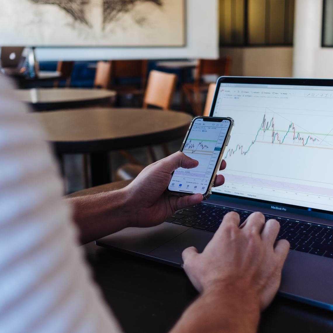 Person holding phone with stock graphs on screen while also on laptop with stock graphs on screen