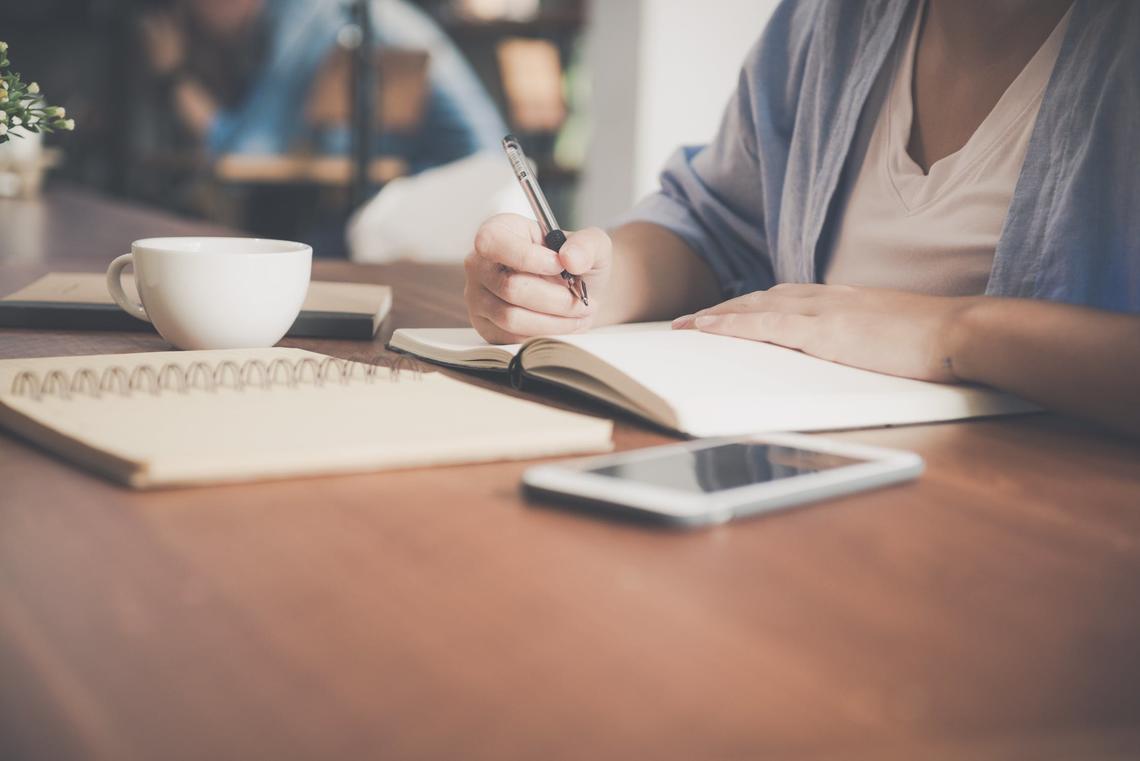 Woman writing on a notebook beside a teacup and tablet