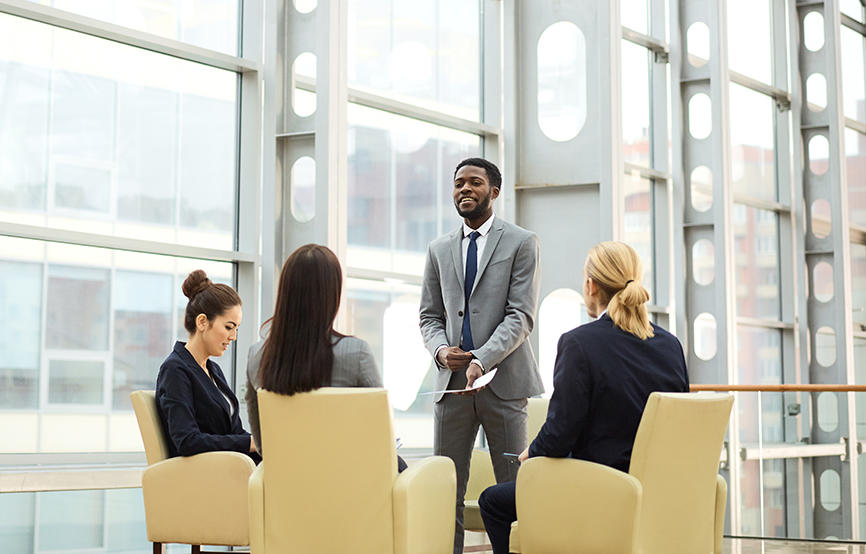 Three people sit in yellow chairs, looking at a person speaking, in a bright, well-lit space