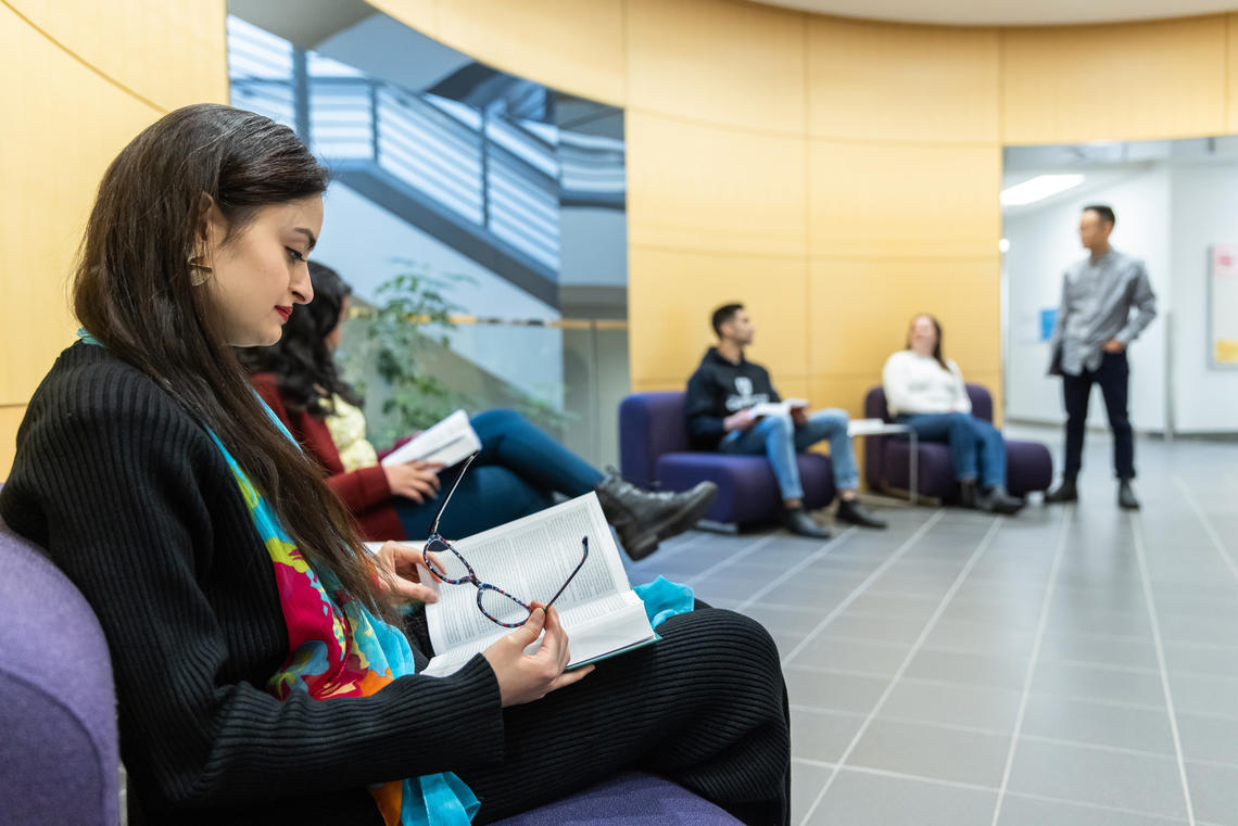 A student reads a book in a common area. In the background, three students are talking. 