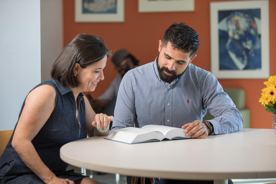 Two students sit at a table looking at a textbook