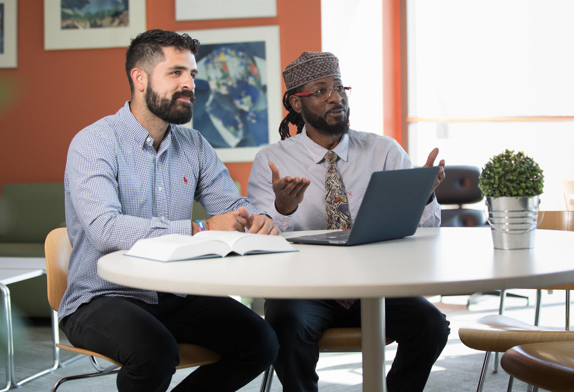 Two students sit at a table with a laptop and textbook in front of them