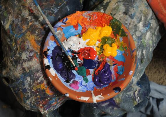 A brown bowl is filled with different colours of paint. A blue paintbrush rests in the bool, which is sitting on a paint splattered drop cloth.