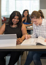 Two students sit at a table, with other students in the background. One has a tablet in front of them and the other is looking in a textbook.