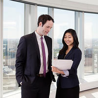 A man and woman stand in front of a window overlooking Calgary. They are looking at a stack of papers held by the woman.