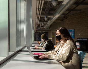 Students sit at a long desk in front of a window working on laptop computers. They are wearing masks.