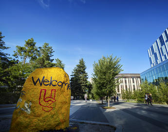 A shot of campus on a sunny day. There is a large rock in the foreground that is painted yellow with the word "Welcome."