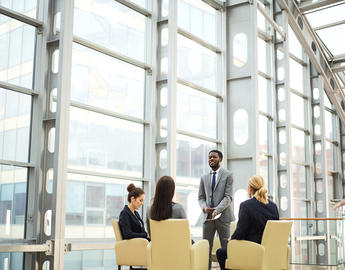 Group of individuals in business wear, with three sitting down and one standing and speaking. 