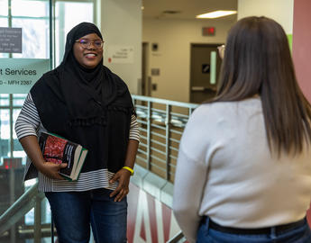 Two students stand in a hallway talking. One is holding books and wearing a hijab.