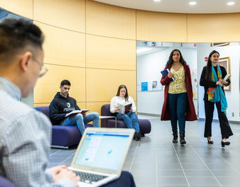 Two students walk through a hallway, while three students are seated and studying.