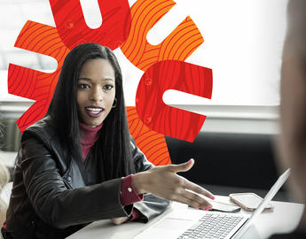 A student sits at a table with a laptop in front of her. She is talking and gesturing to a student across from her.